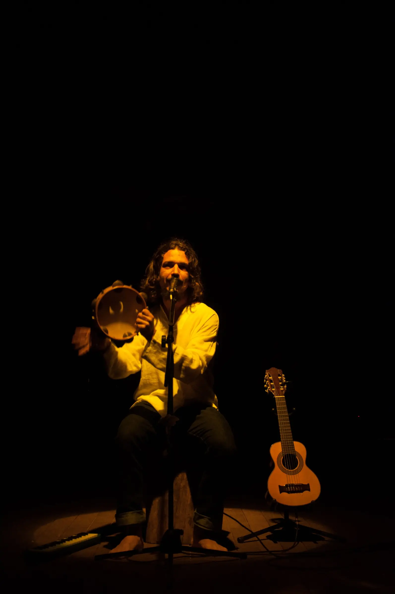 Rodolfo Minari no palco, sob luz dourada, tocando pandeiro ao lado de um violão